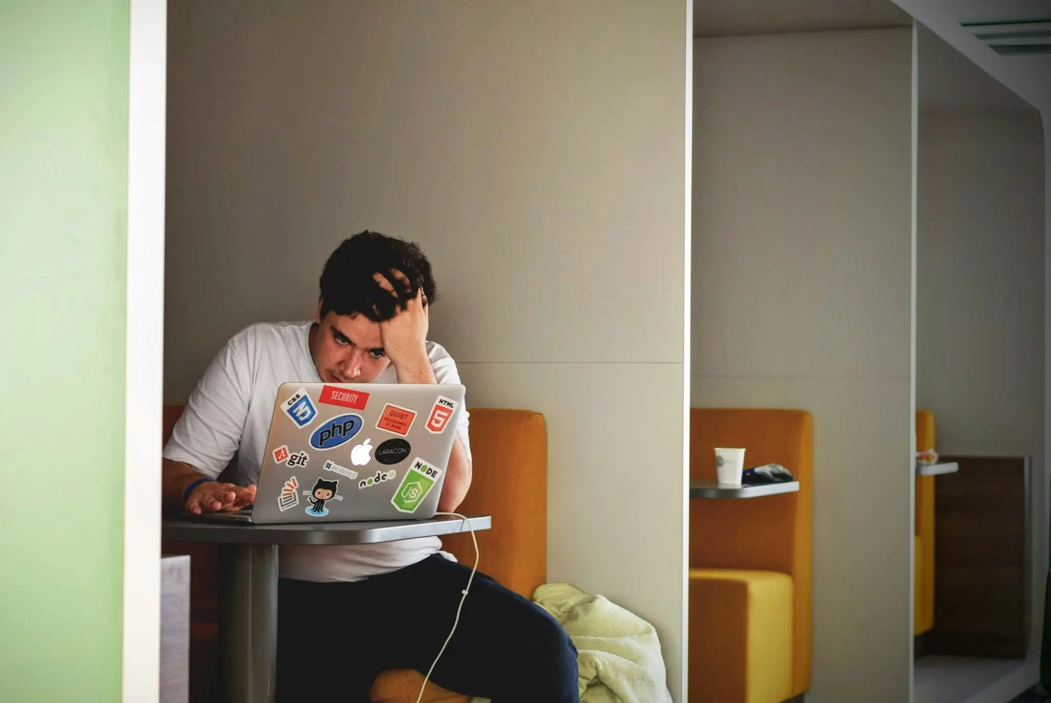 A young man sitting in a coffee shop booth alone, looking at his laptop screen with a confused and stressful expression.